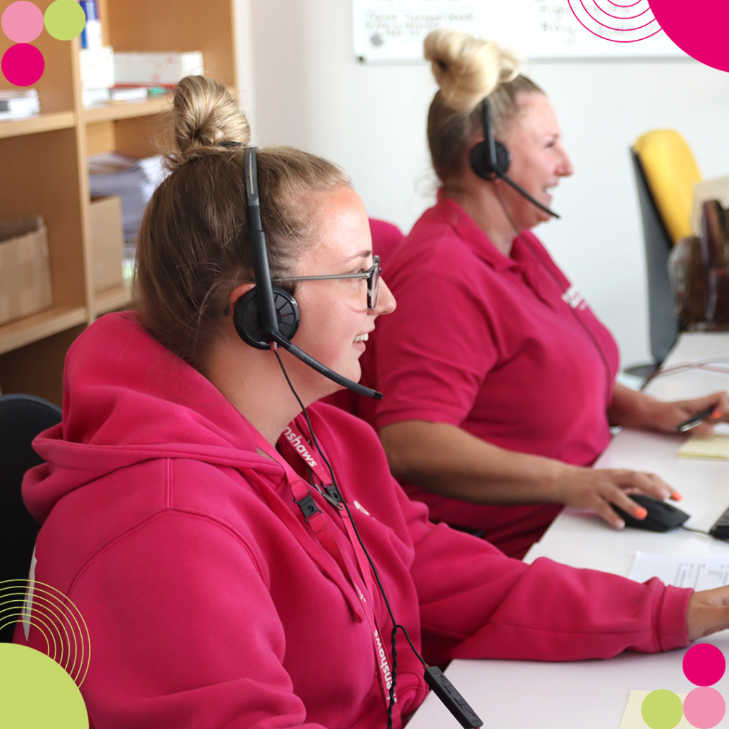 Two women with blonde hair, wearing pink clothing, they are sitting at a desk. They are both wearing headsets with microphones.