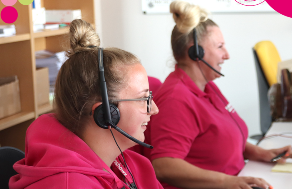 Two women with blonde hair, wearing pink clothing, they are sitting at a desk. They are both wearing headsets with microphones.