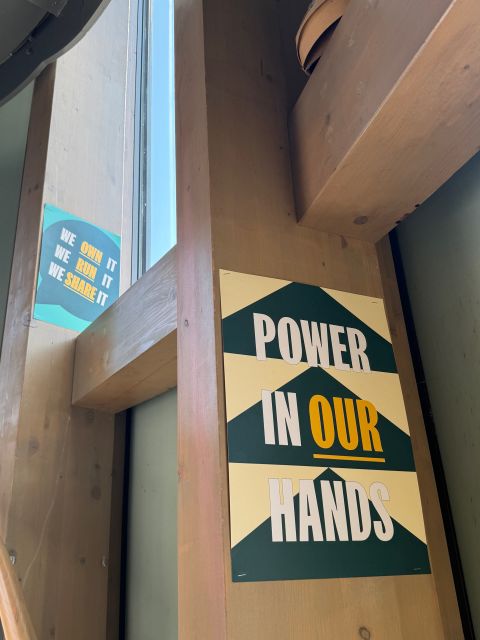 Poster reading ‘Power in Our Hands’ displayed on a wooden pillar inside a community venue, alongside a smaller sign that reads ‘We own it, we run it, we share it.’
