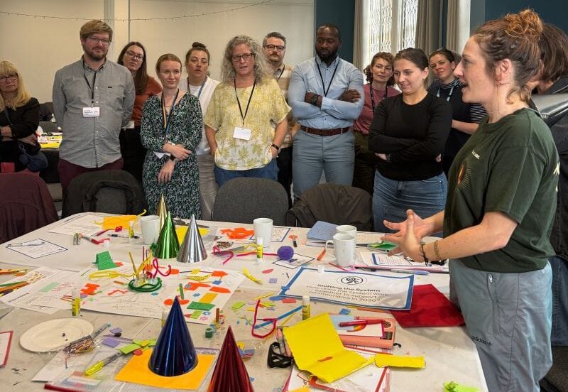 A group of around a dozen people stand around a large table covered with craft materials, sticky notes, pens and paper during a workshop activity. One person stands at the front speaking to the group while others gather around and listen. The table contains colourful materials, including pipe cleaners, card, glue sticks and cone-shaped objects, suggesting a creative or collaborative session in a meeting room.