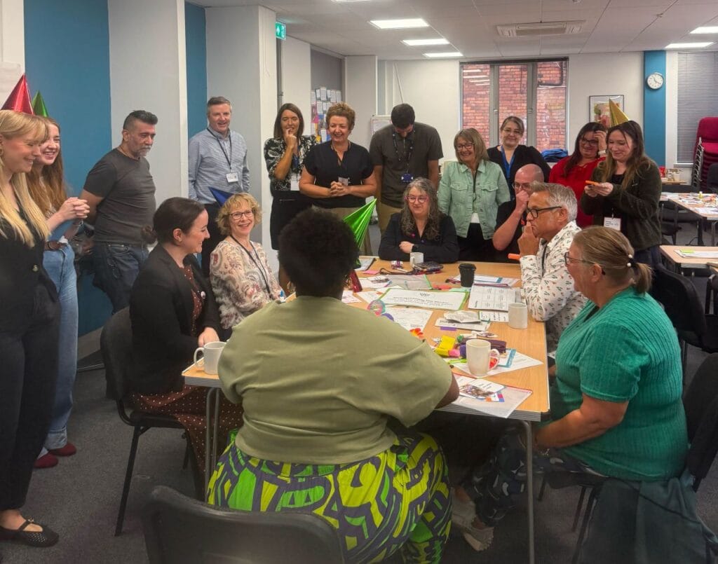 A group of people gather around a table during a workshop session in a meeting room. Several people are seated at the table, which is covered with worksheets, stationery, mugs and colourful craft materials, while others stand around observing and taking part in the discussion. Some participants wear party hats, creating a relaxed and creative atmosphere. The room has large windows, office furniture and workshop resources visible in the background.