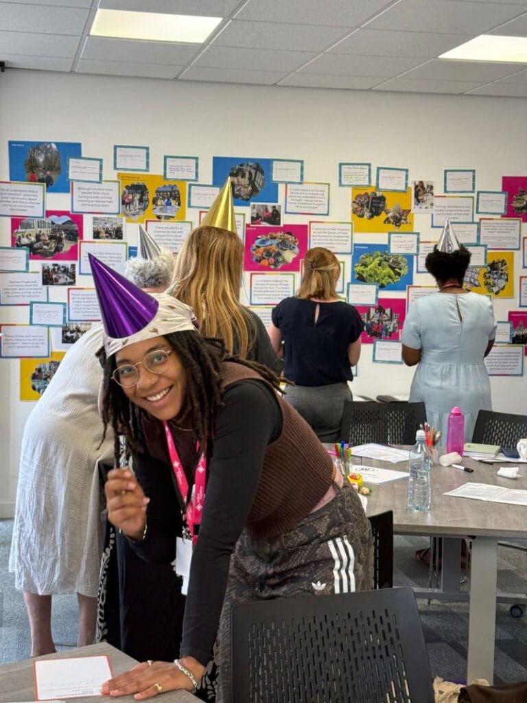 Participants stand around a table covered with notes, pens and craft materials, listening to a group discussion during a collaborative workshop