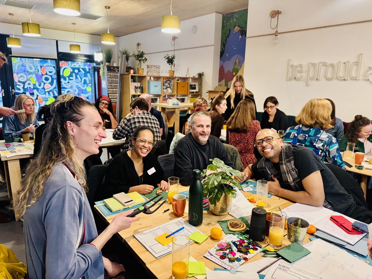 A group of around 15 people sit in a workshop at the Proud Trust community café. Notebooks, papers, sticky notes, drinks and snacks are spread across the tables. Several participants are engaged in discussion while others listen or write notes. Shelving, plants and colourful artwork are visible in the background, creating an informal and welcoming workshop setting.