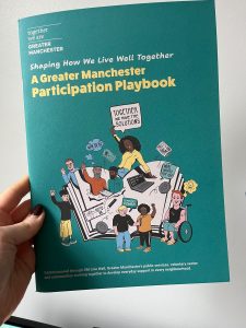 A person holds a copy of the &ldquo;Greater Manchester Participation Playbook&rdquo; against a plain wall. The teal cover features illustrations of community members gathered around an open book, speech bubbles with messages including &ldquo;Together we have the solutions&rdquo;, and imagery representing participation, community power and shared decision-making.